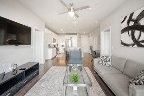 A modern living room with a flat-screen TV, a glass coffee table, a sofa, and a ceiling fan.