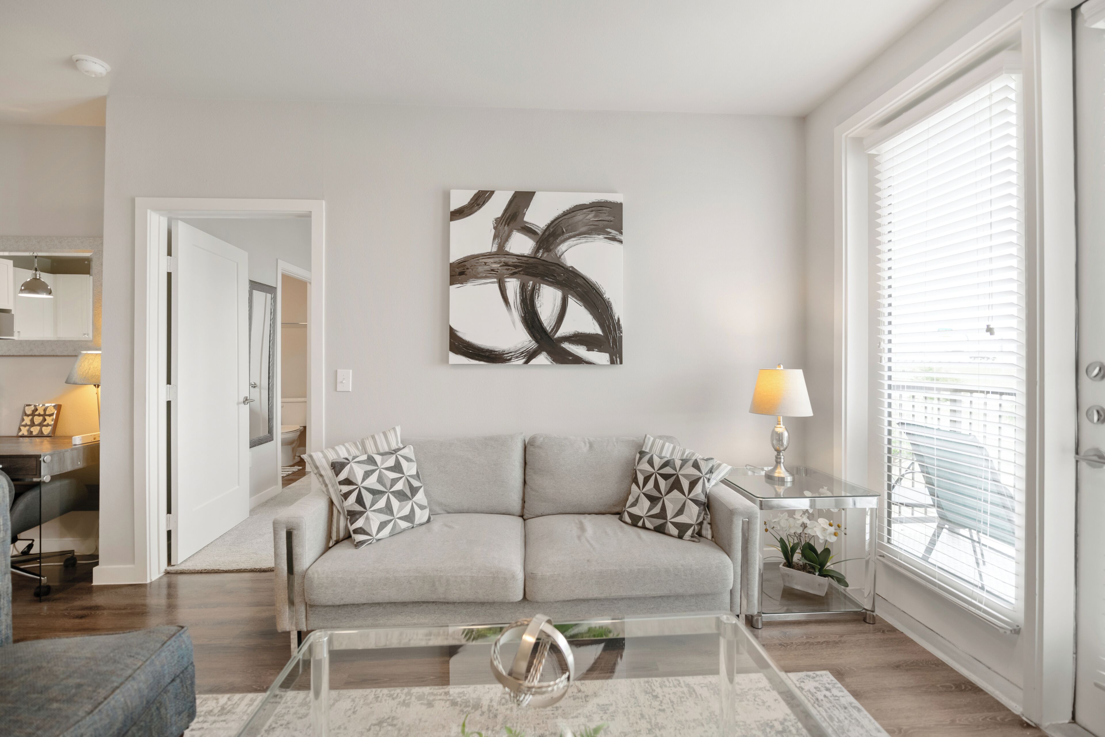 Een moderne woonkamer met een grijze bank, abstracte kunst, een glazen salontafel en een uitzicht door witte jaloezieën.