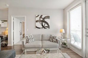 A modern living room with a grey sofa, abstract art, a glass coffee table, and a view through white blinds. - MCK211 | McKinney Corporate 2BD | Pool | Workspace (McKinney)