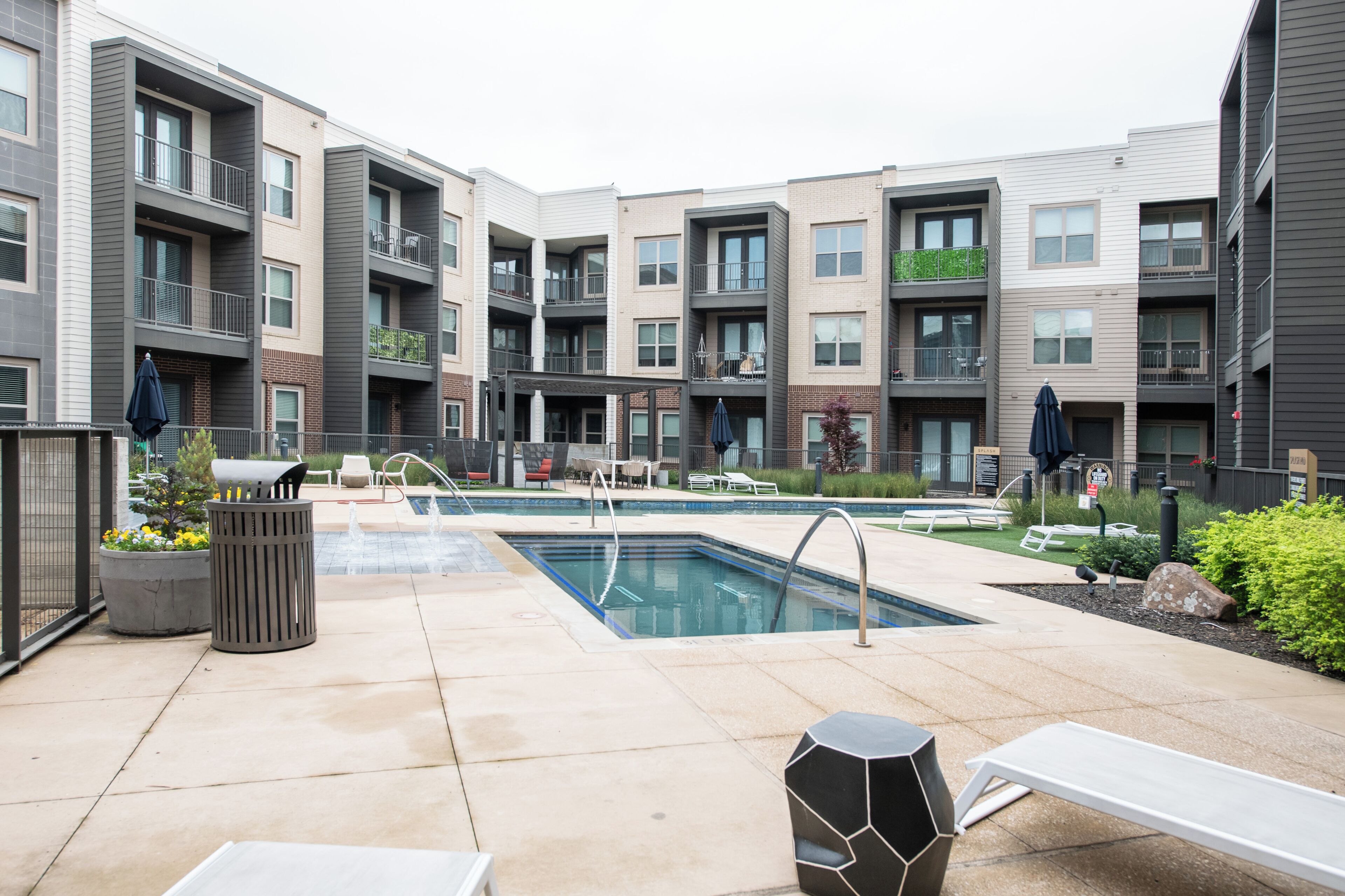 A modern apartment complex with a central swimming pool, outdoor seating, and a landscaped garden area.