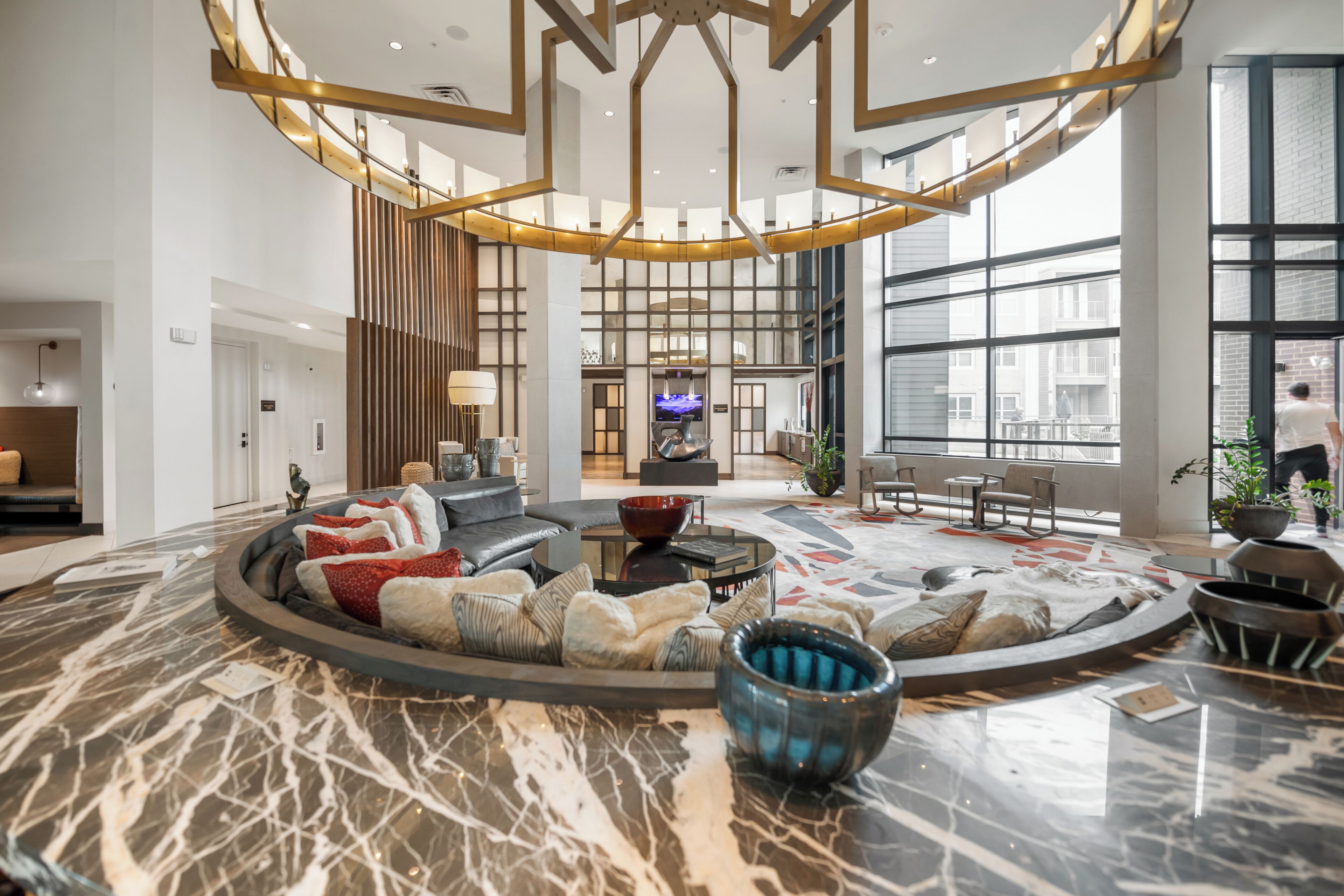 A spacious lobby with a central seating area, a large window offering a view of the city, and a modern chandelier.