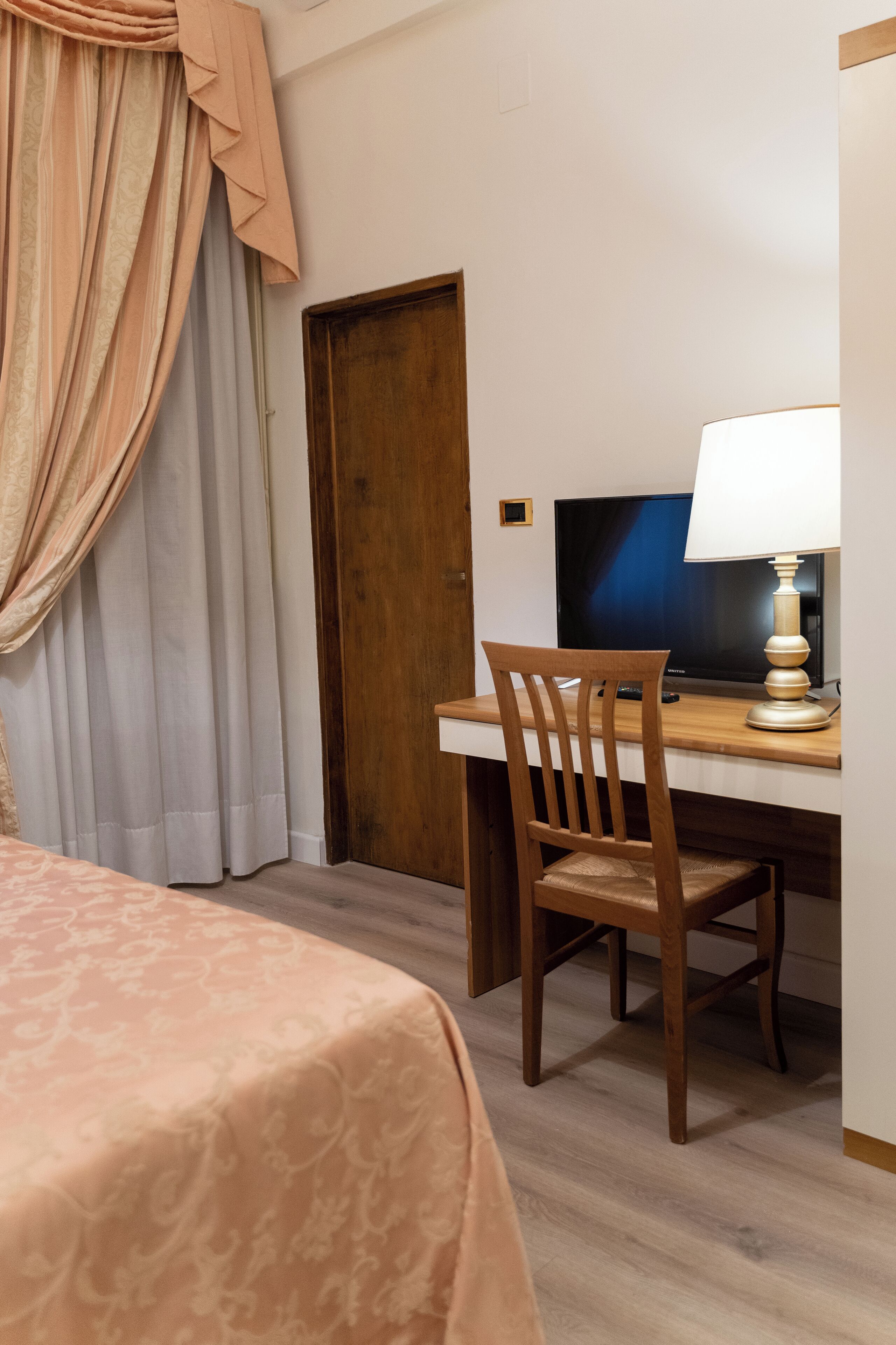 A hotel room with a bed, a wooden desk with a computer and lamp, and a wooden chair.