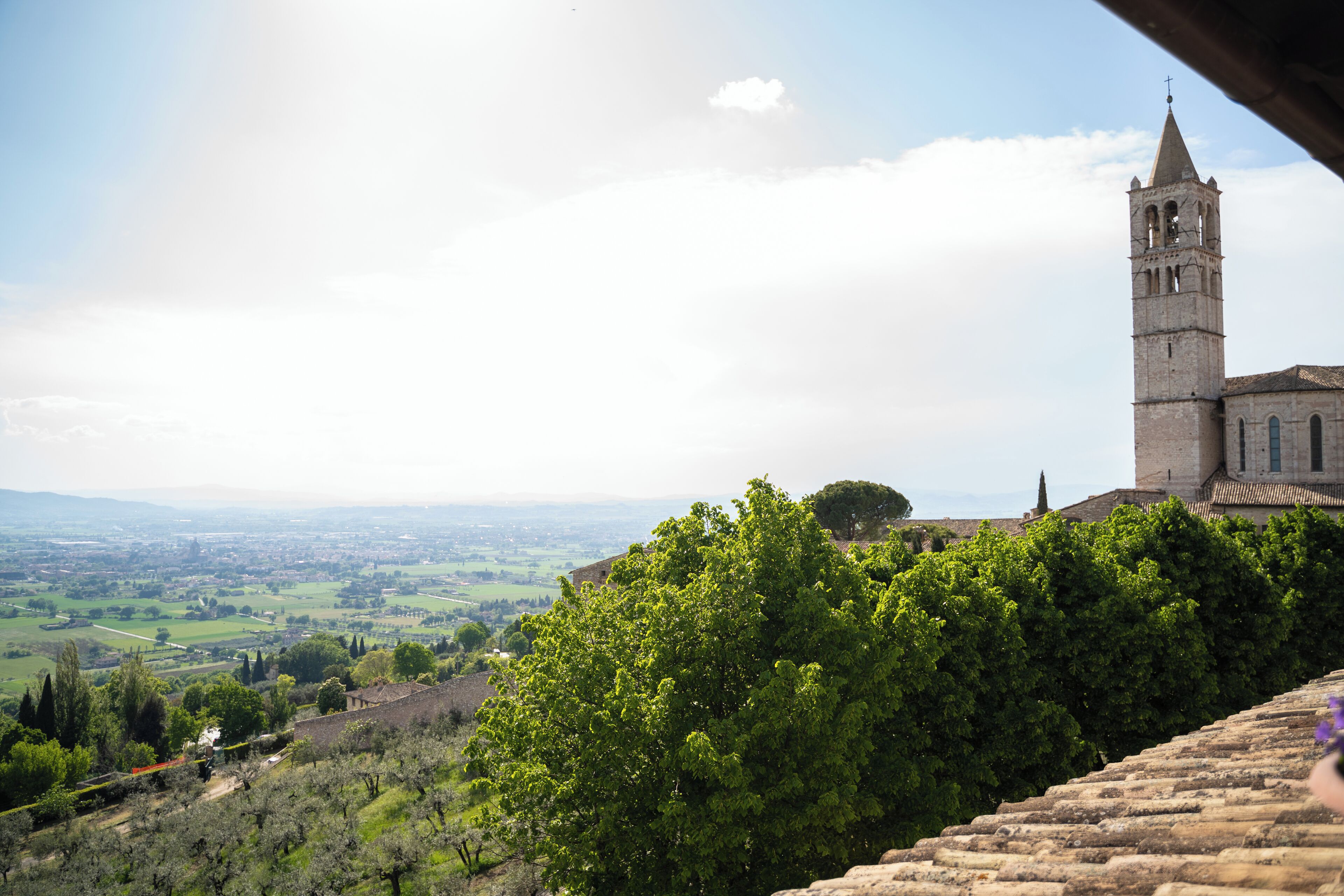 A historic stone building with a bell tower, surrounded by lush greenery and a view of a distant valley.