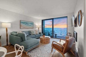 A modern living room with a sofa, coffee table, and chairs, a large window with a view of the sea, and a flat-screen TV mounted on the wall.