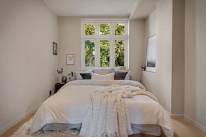 A bedroom with a large bed, a window with a view of trees, a bedside table with a vase, and a framed picture on the wall.