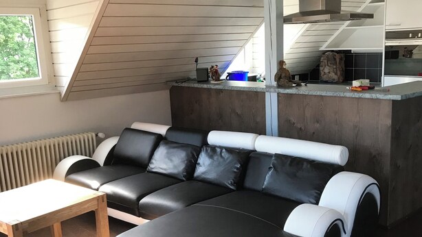 Un espace de vie moderne de style loft, avec un canapé d’angle noir et blanc, une table basse en bois et un coin cuisine doté d’une grande fenêtre.