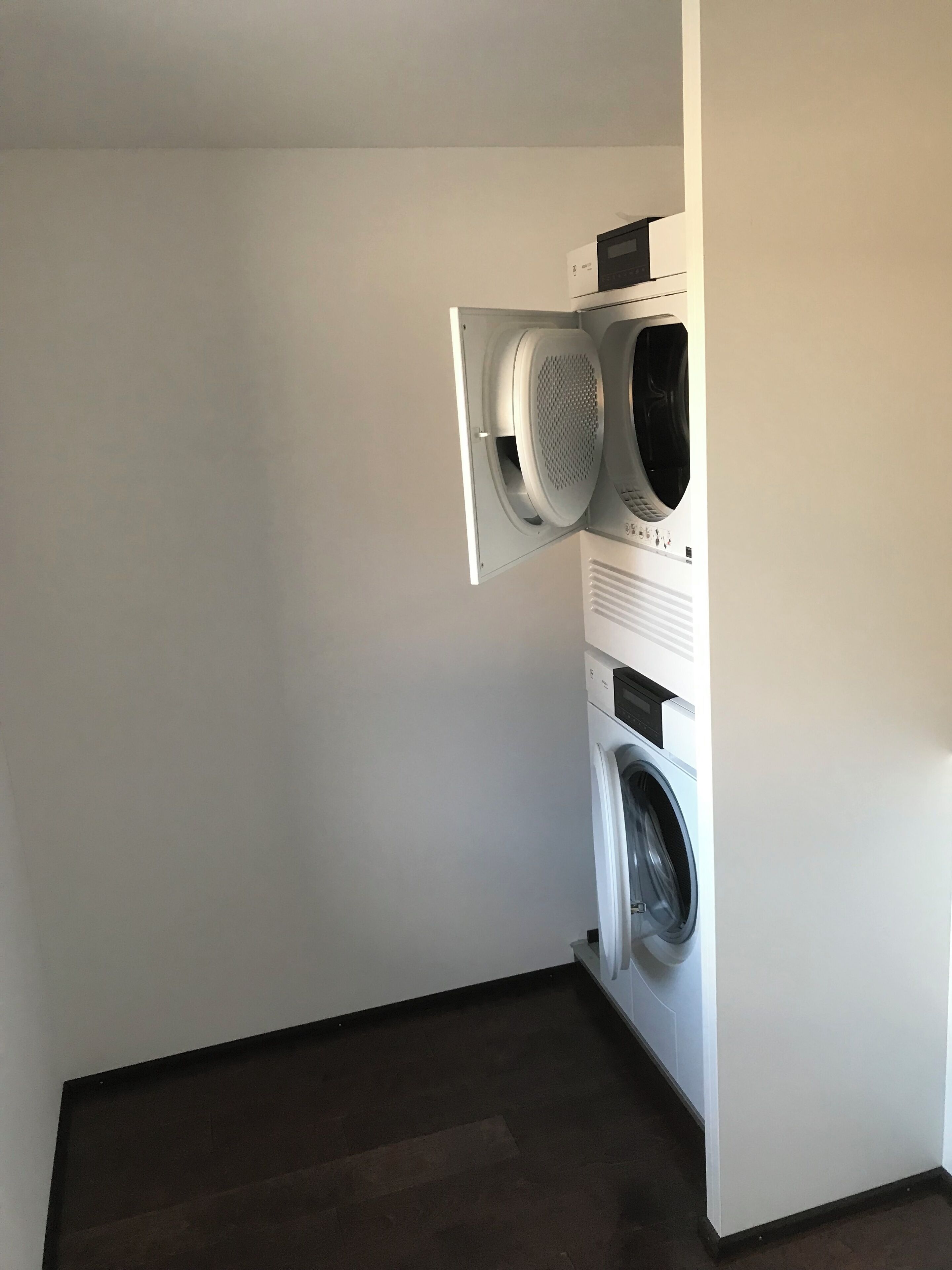 Ein Waschraum mit einer gestapelten Waschmaschine und einem Trockner, einer weißen Wand und einem dunklen Boden.