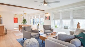 Ein geräumiges Wohnzimmer mit Deckenventilator, einem Essbereich mit Holztisch und -stühlen sowie großen Fenstern mit Jalousien.