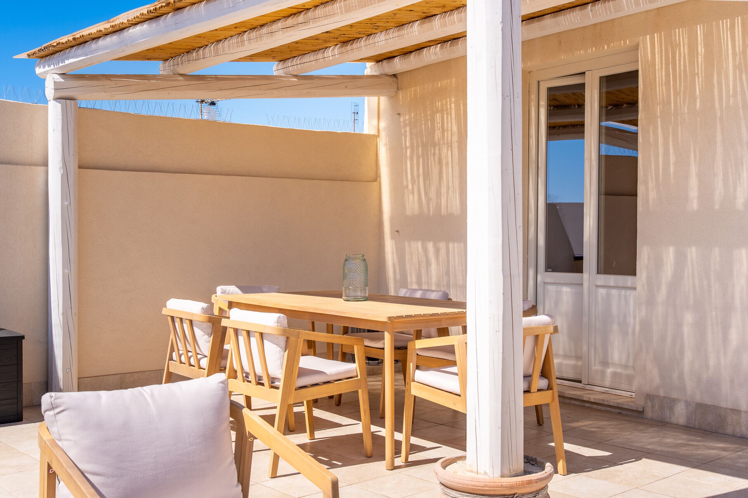 A covered outdoor seating area with a wooden table and chairs, a potted plant, and a door leading to an interior space.