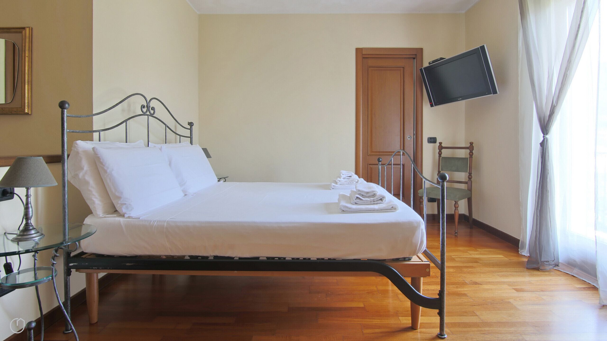 Una cama bien hecha con sábanas blancas, una mesita de noche con lámpara, un televisor montado en la pared y una silla.