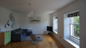Un saló modern amb un sofà gris, un armari gris, un televisor de pantalla plana i un finestral gran amb vista a arbres.