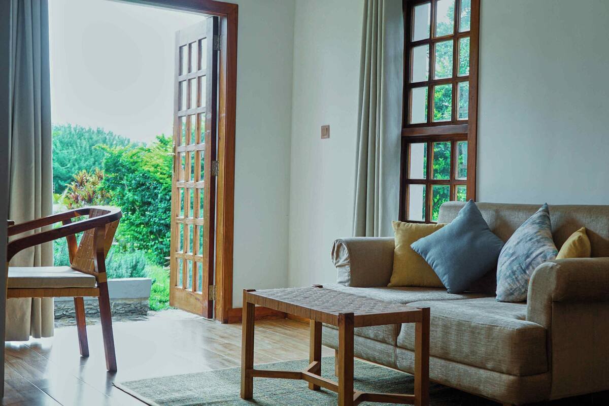 A living room with a sofa, a wooden table, and a chair. There is a large window with curtains and a view of greenery outside.