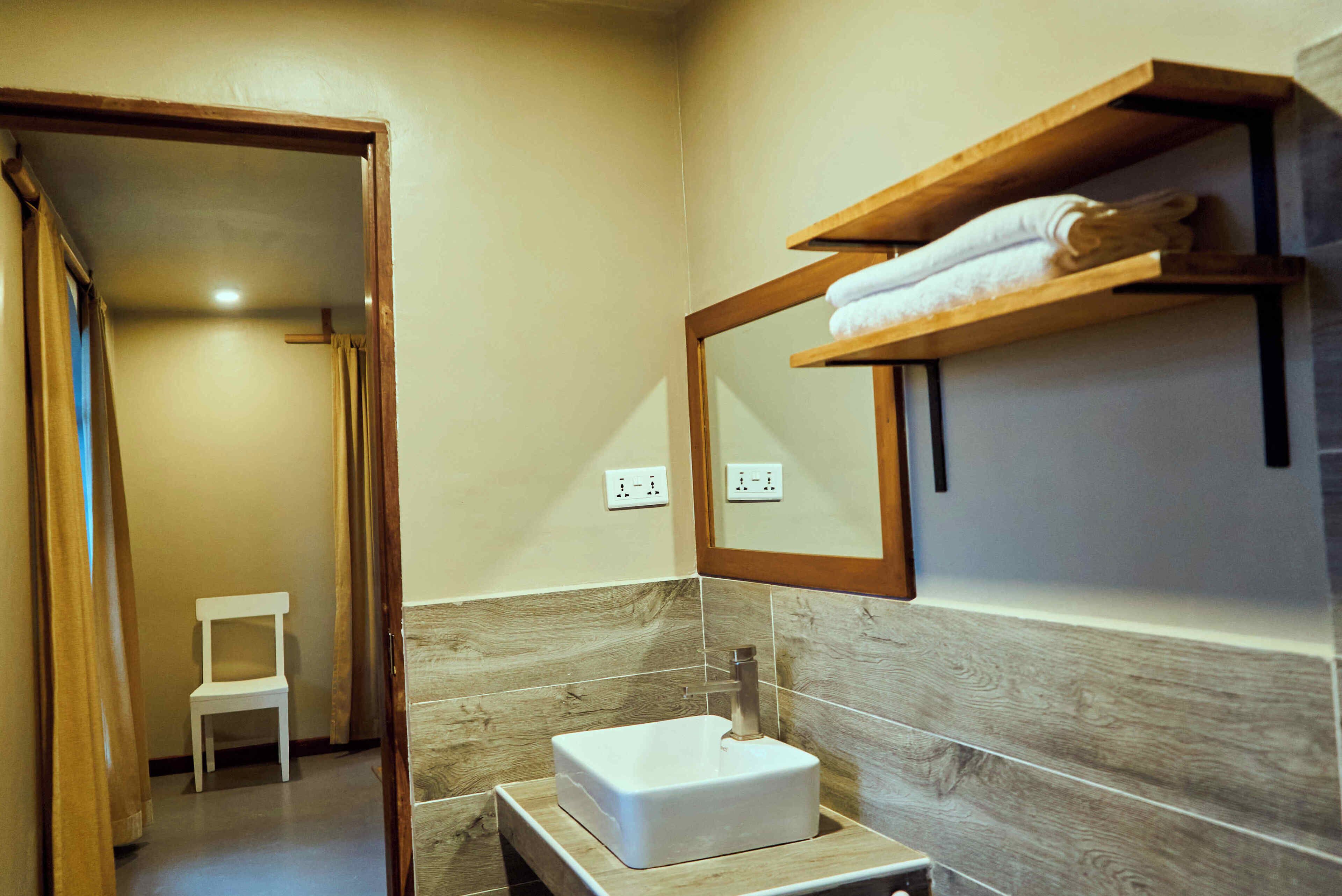 A bathroom with a white sink, wooden shelves, a mirror, and a chair.