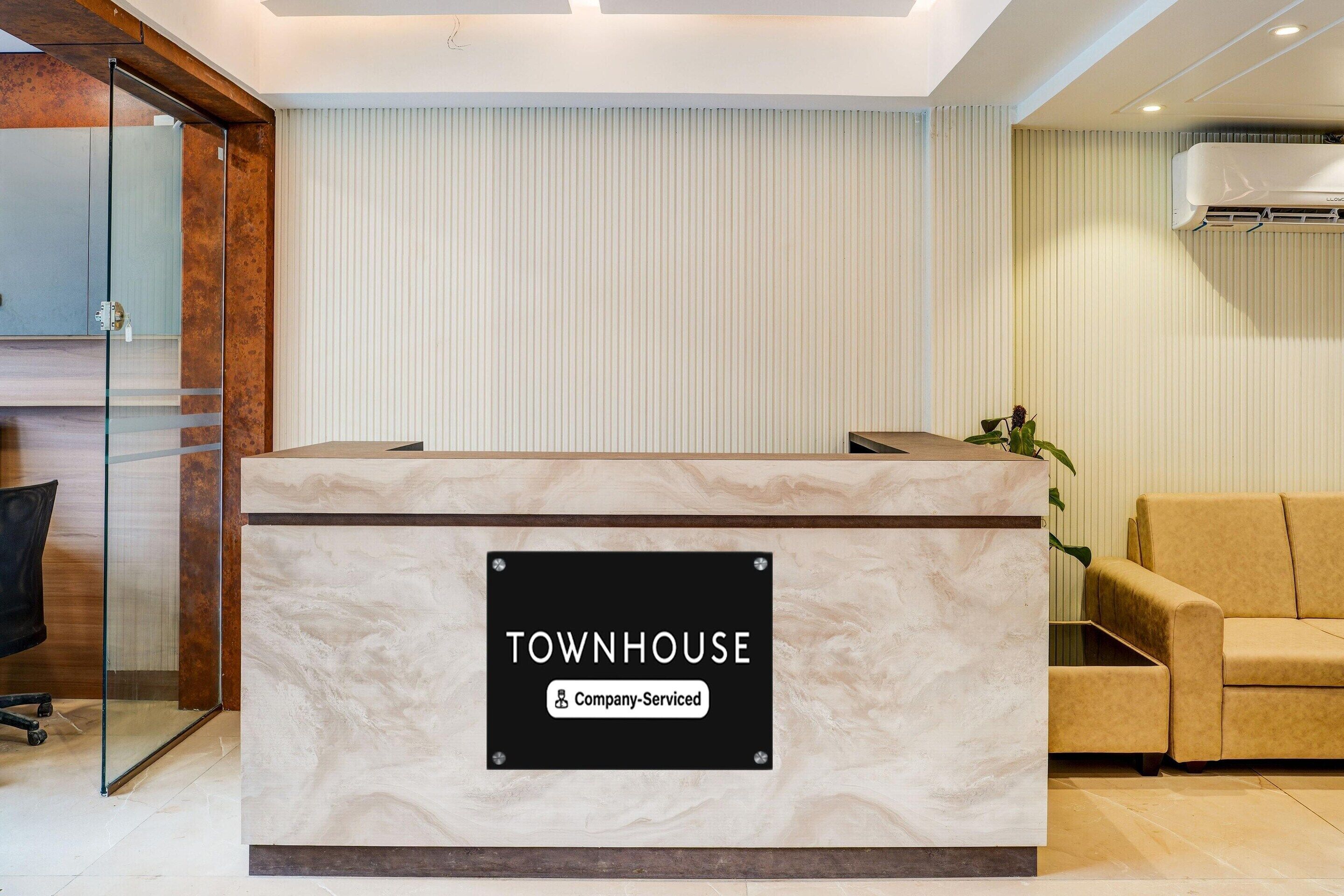 Ένας πάγκος υποδοχής με την επιγραφή «TOWNHOUSE» και μια πινακίδα που αναγράφει «Υπηρεσίες Εταιρείας», ένα γυάλινο διαχωριστικό, ένας μπεζ καναπές και μια μονάδα κλιματισμού στον τοίχο.