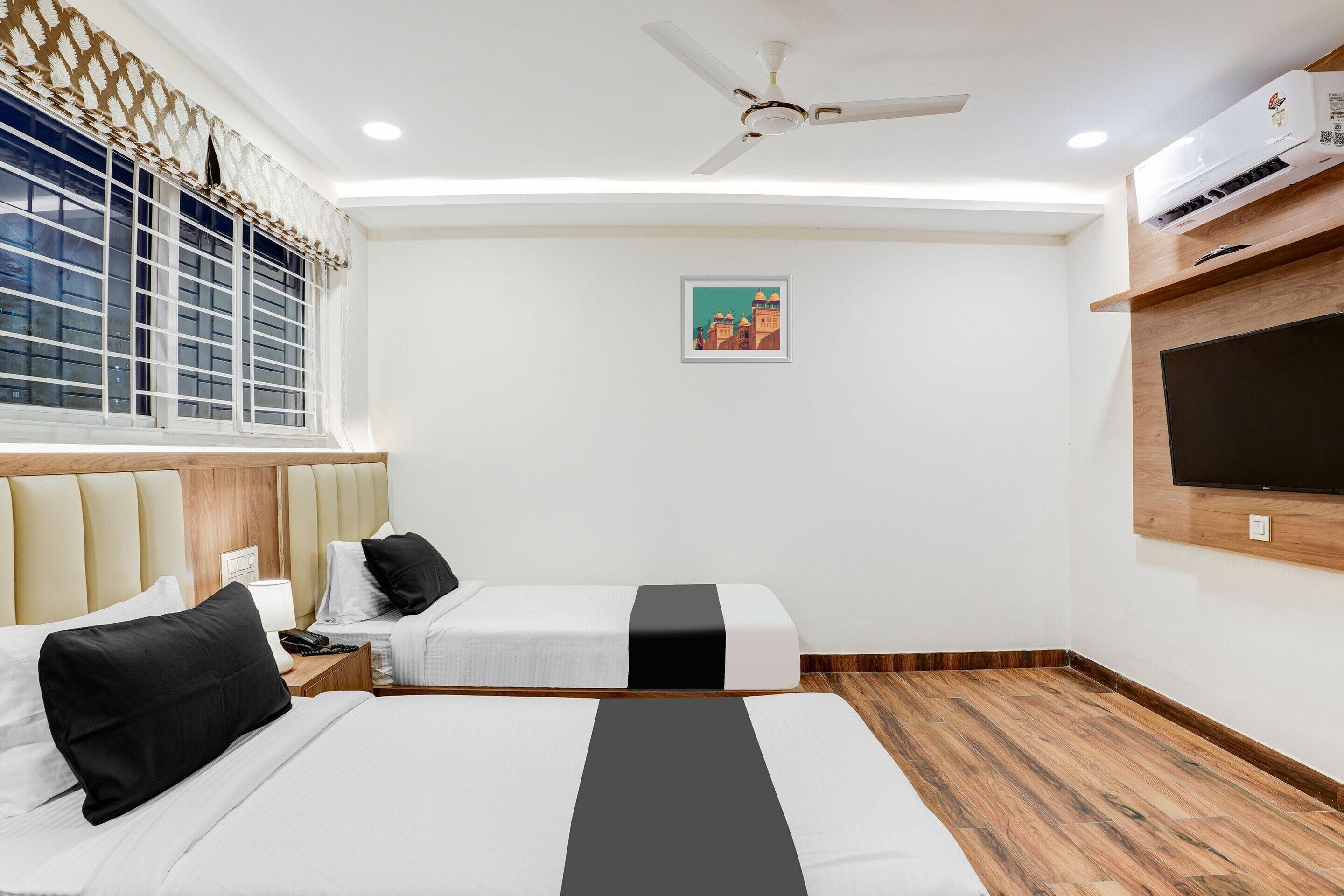 A hotel room with two beds, a ceiling fan, an air conditioning unit, a mounted TV, and a window with decorative curtains.