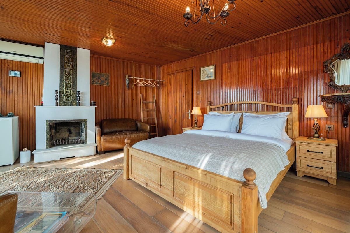 Quarto com painéis de madeira, uma cama grande, lareira, cadeira e mesa de cabeceira com abajur.