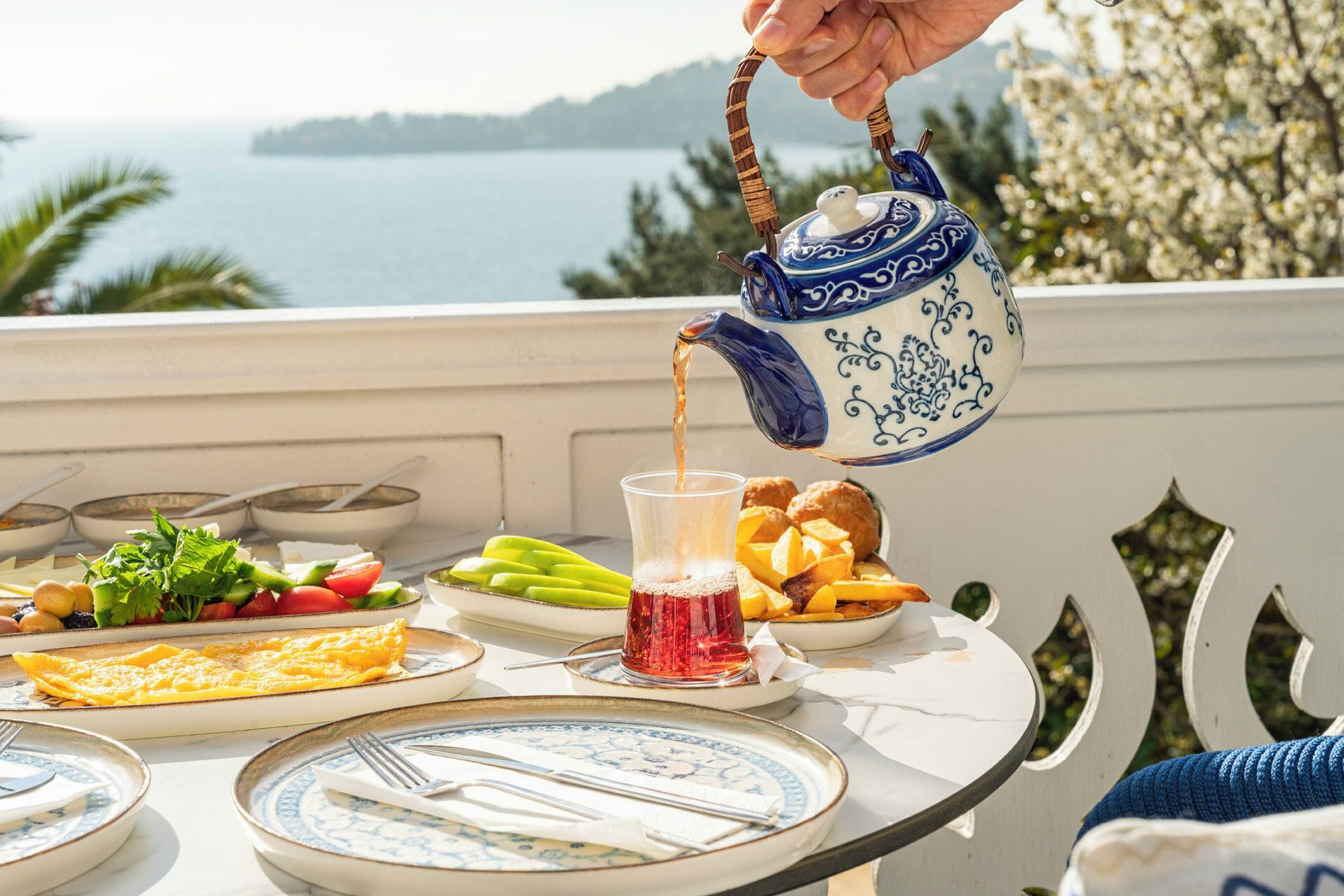 A table set with breakfast items, including eggs, fruits, and pastries, with a scenic view of the sea in the background.