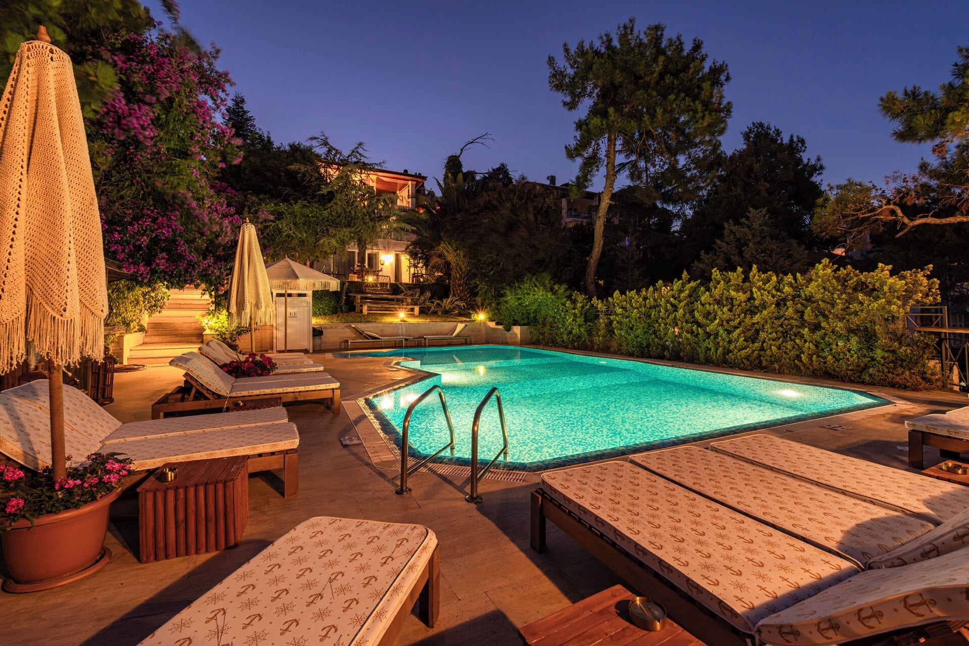 A swimming pool with lounge chairs and umbrellas, surrounded by trees and a building in the background.