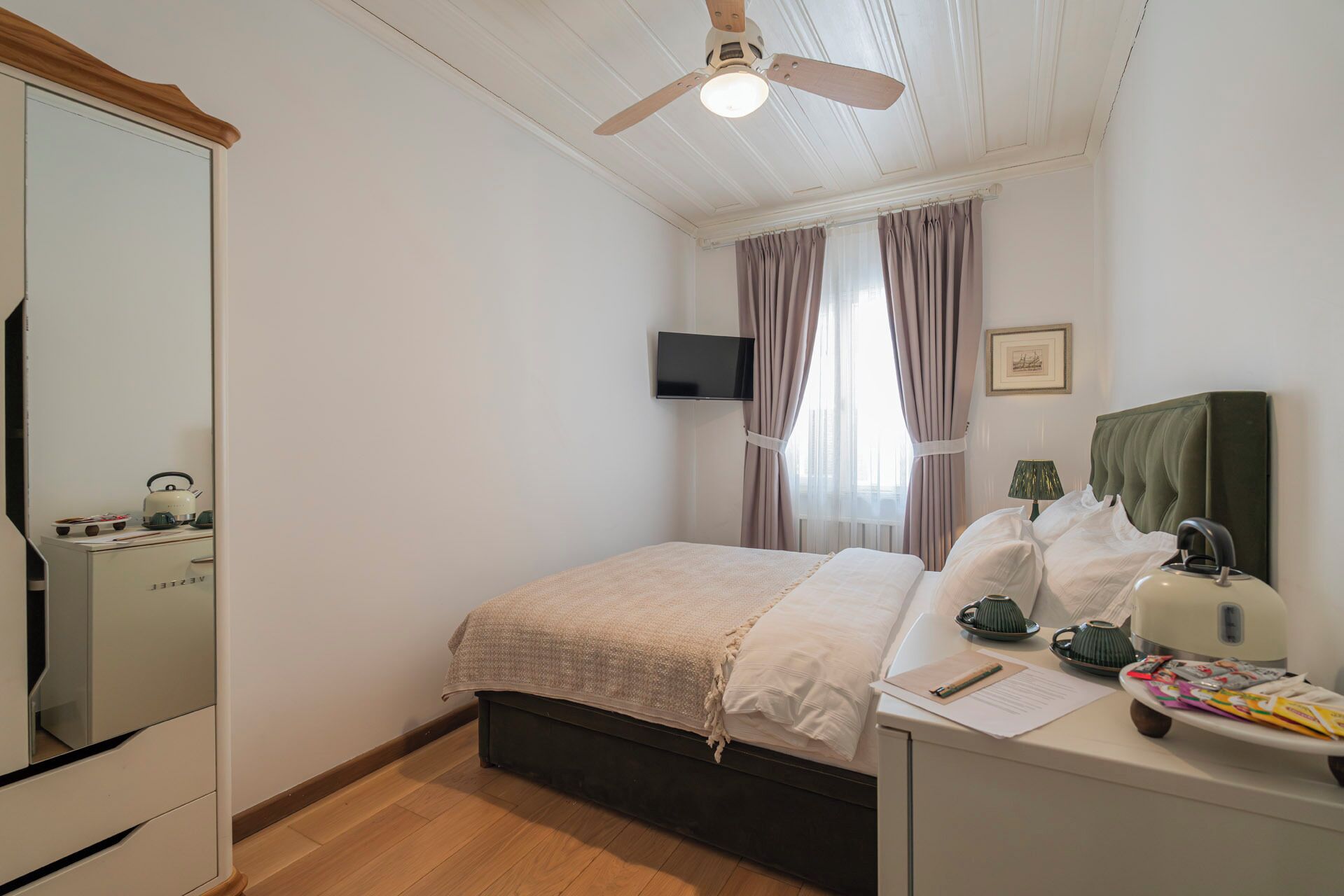 A bedroom with a bed, a bedside table with a lamp, a mirror, a ceiling fan, and a TV on the wall.