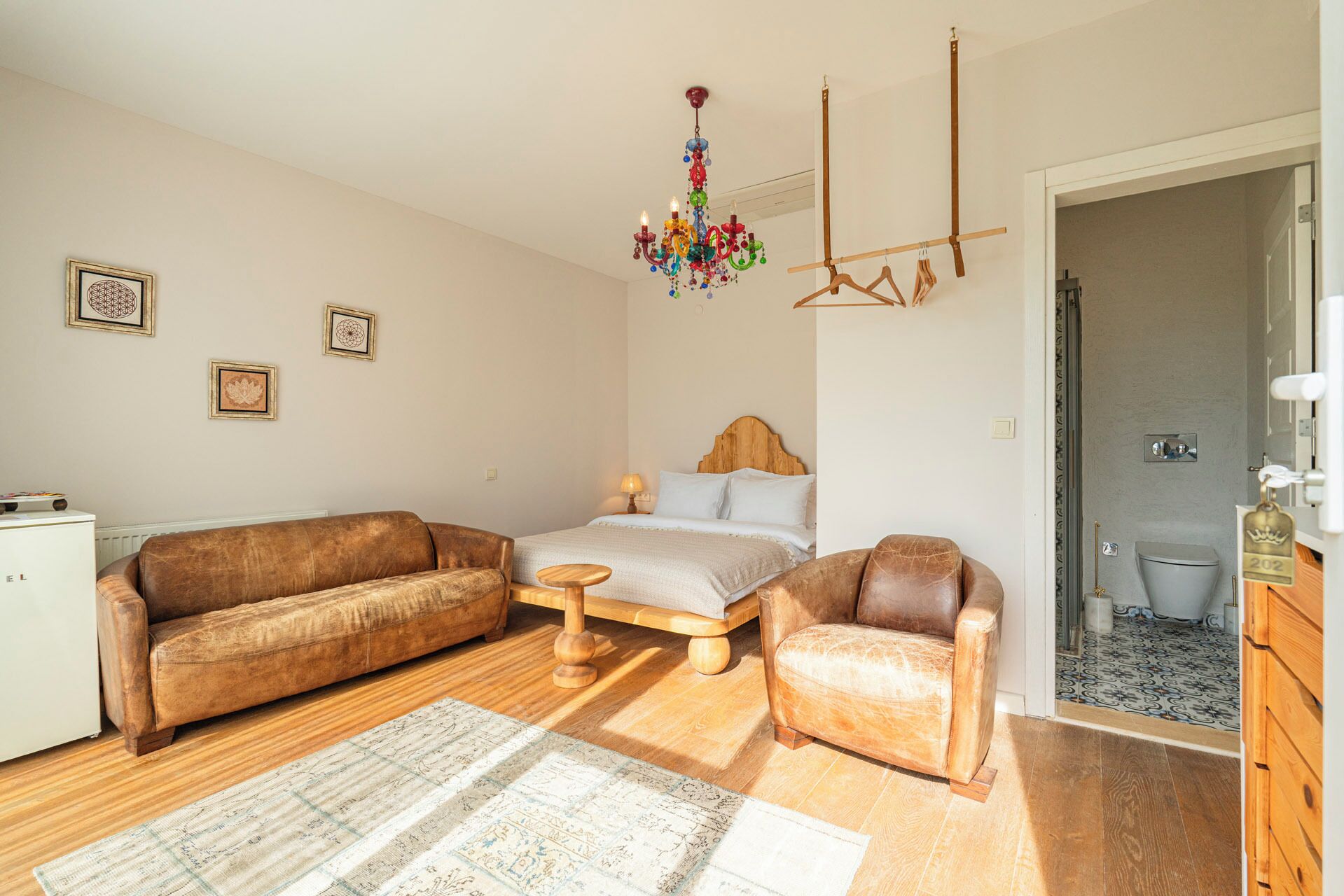 Een knus ingerichte kamer met een bed, twee bruine leren banken, een houten bijzettafeltje, een kleurrijke kroonluchter en een klein witte radiator.