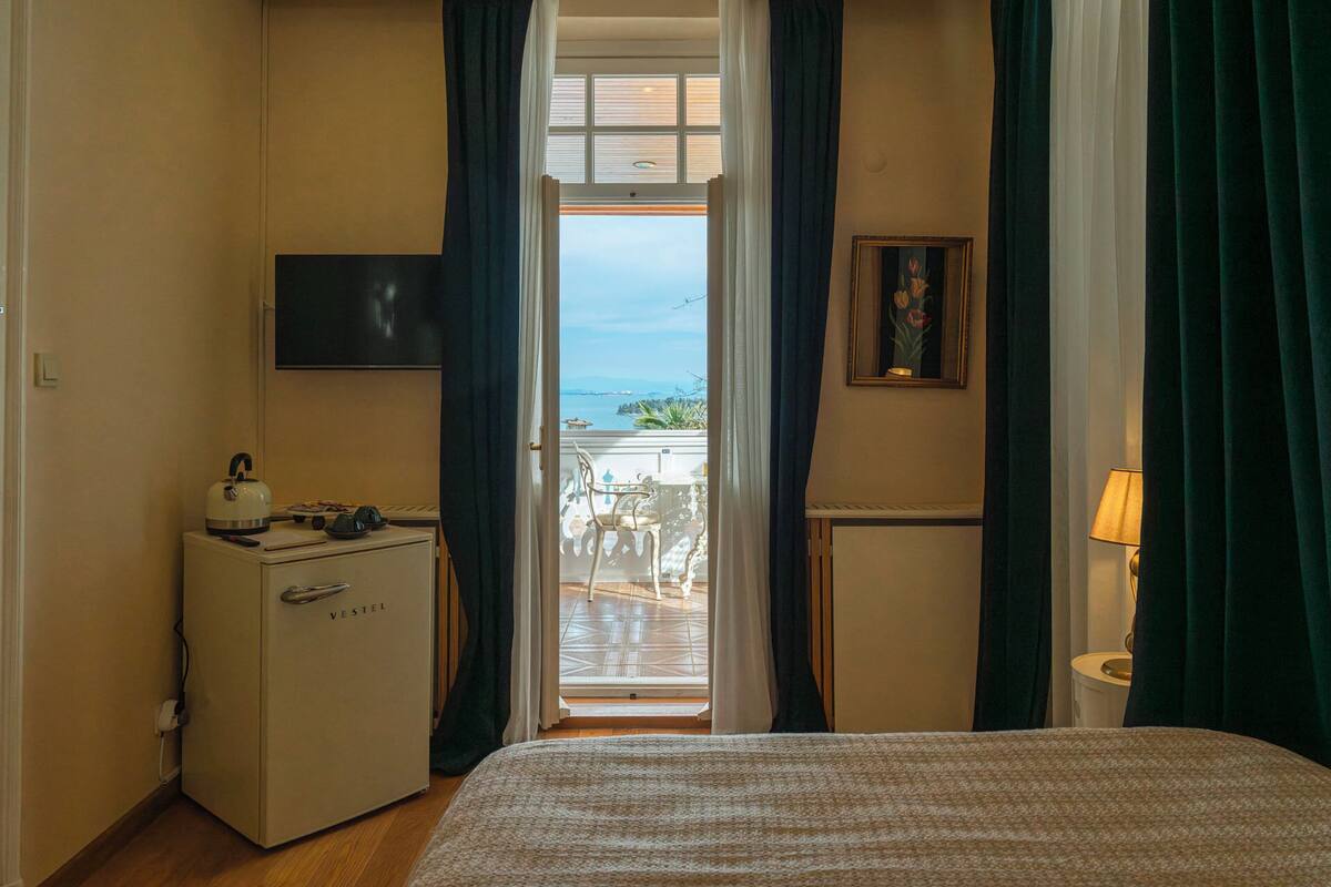 Quarto de hotel com uma cama, escrivaninha com chaleira, televisão, quadro e vista para o mar.