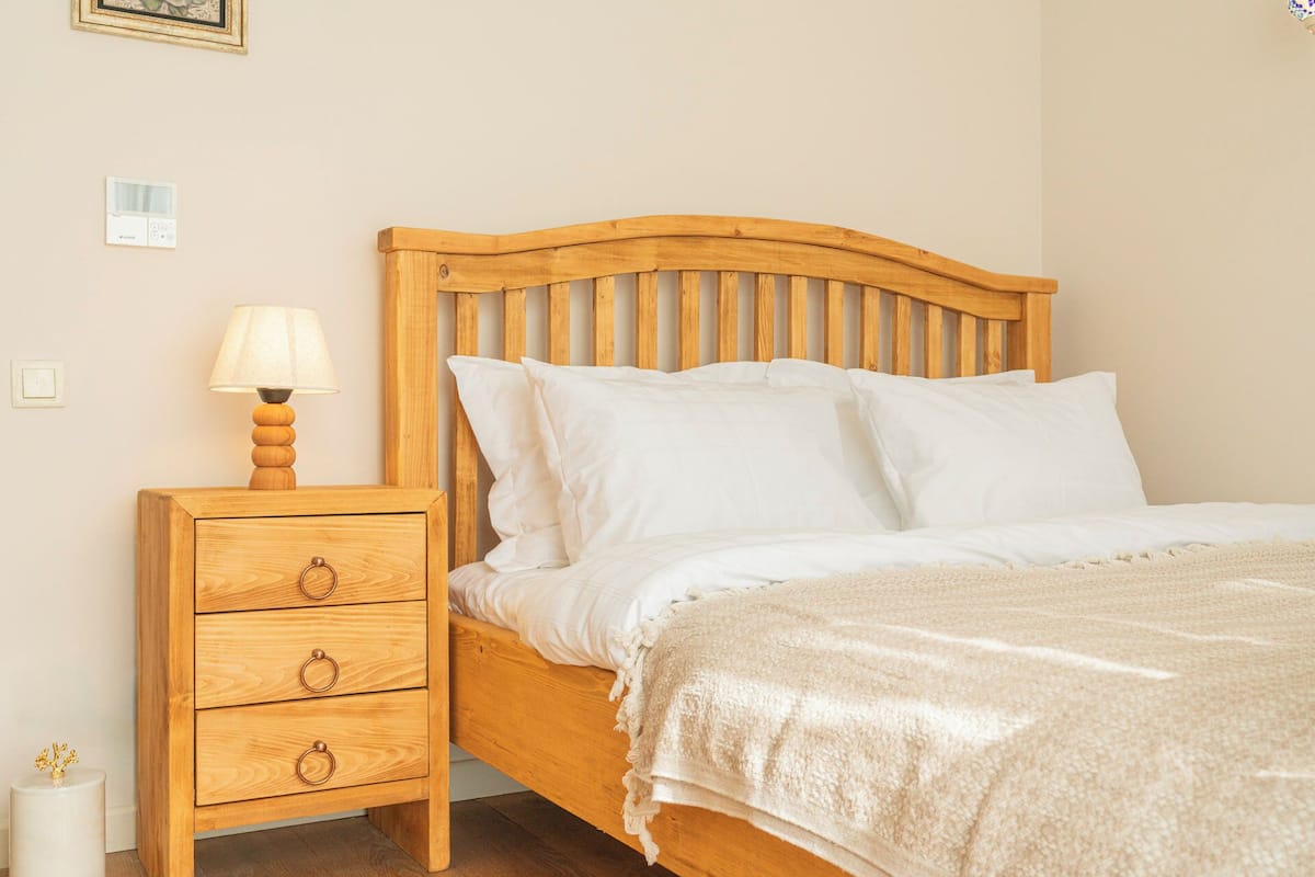 Uma cama de madeira com roupa de cama branca, uma mesa de cabeceira com abajur e uma cômoda.