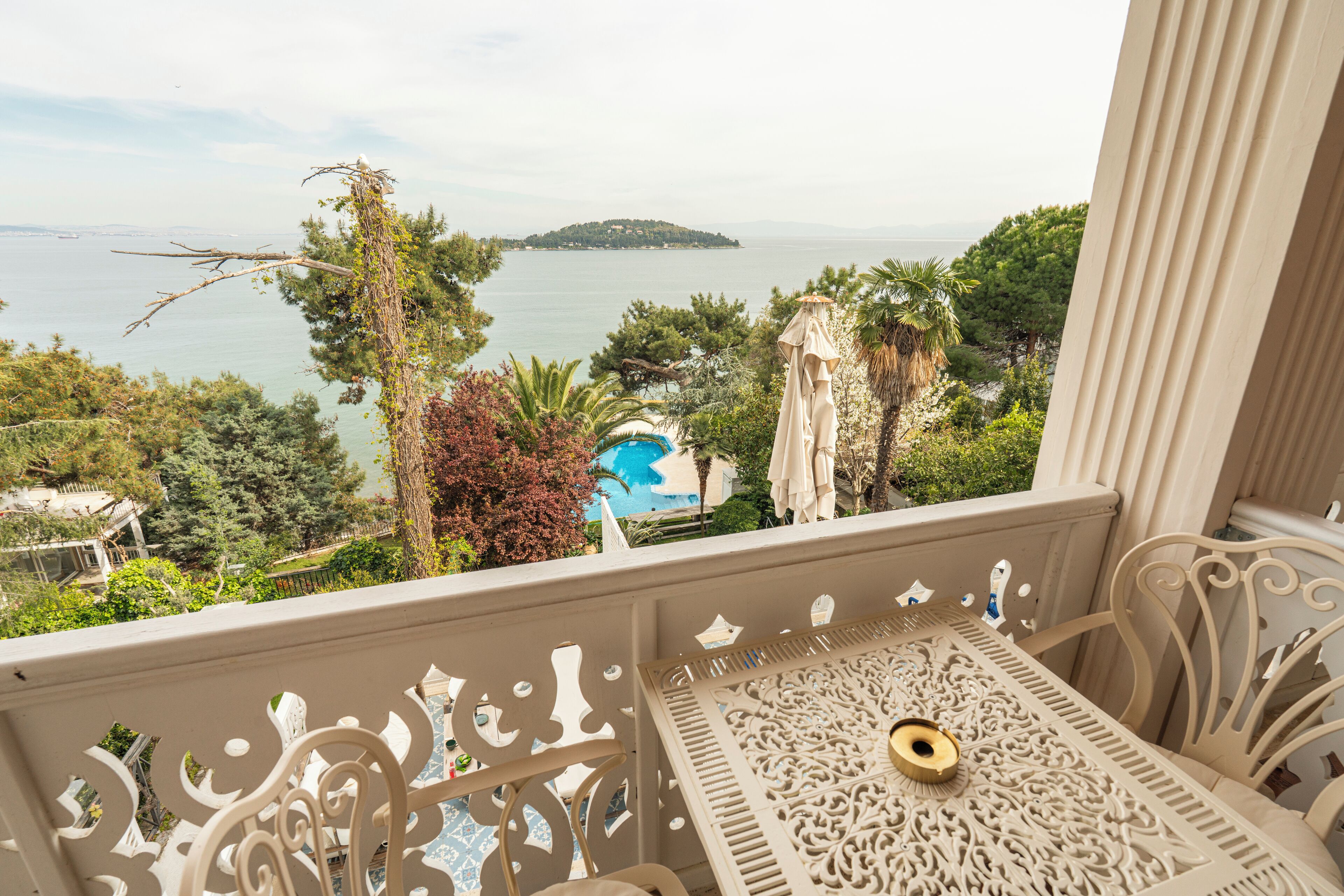 A balcony with ornate metal railings, a table with a decorative centerpiece, and a view of a body of water with a distant island and a pool.