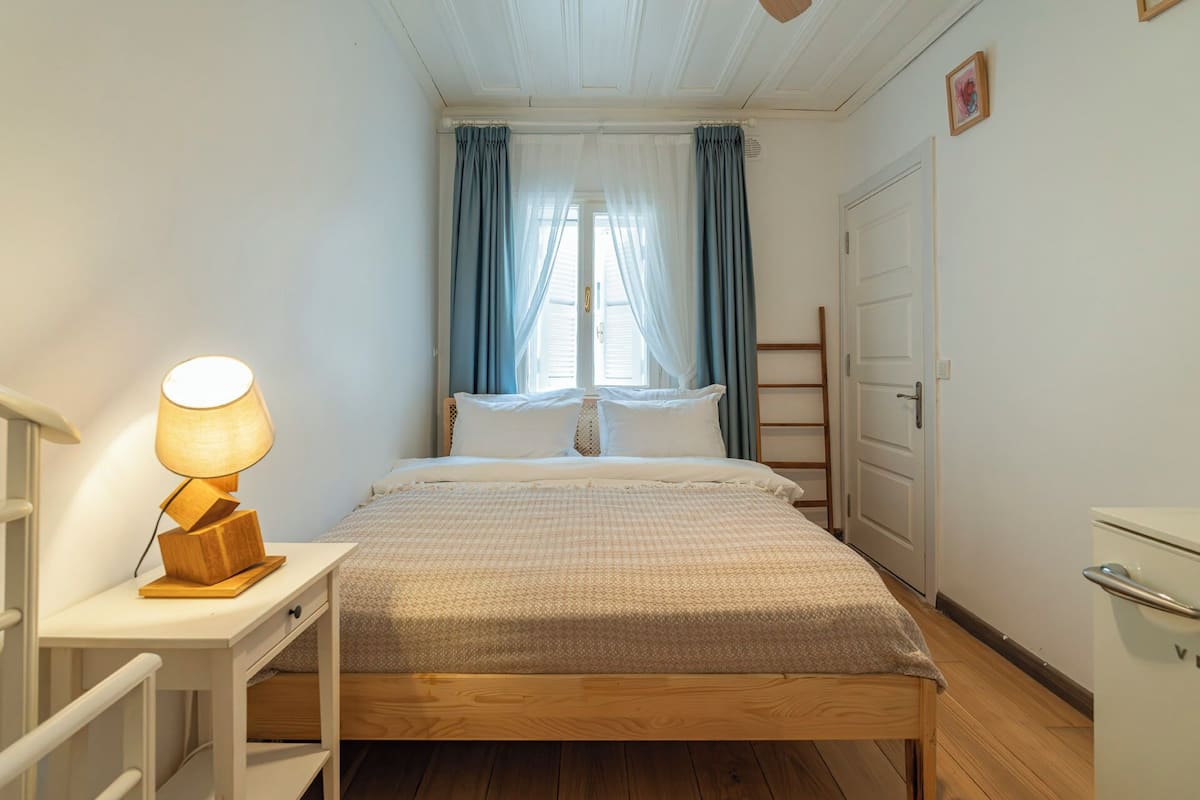 Quarto com cama de madeira, mesa de cabeceira com luminária, janela com cortinas e uma porta.