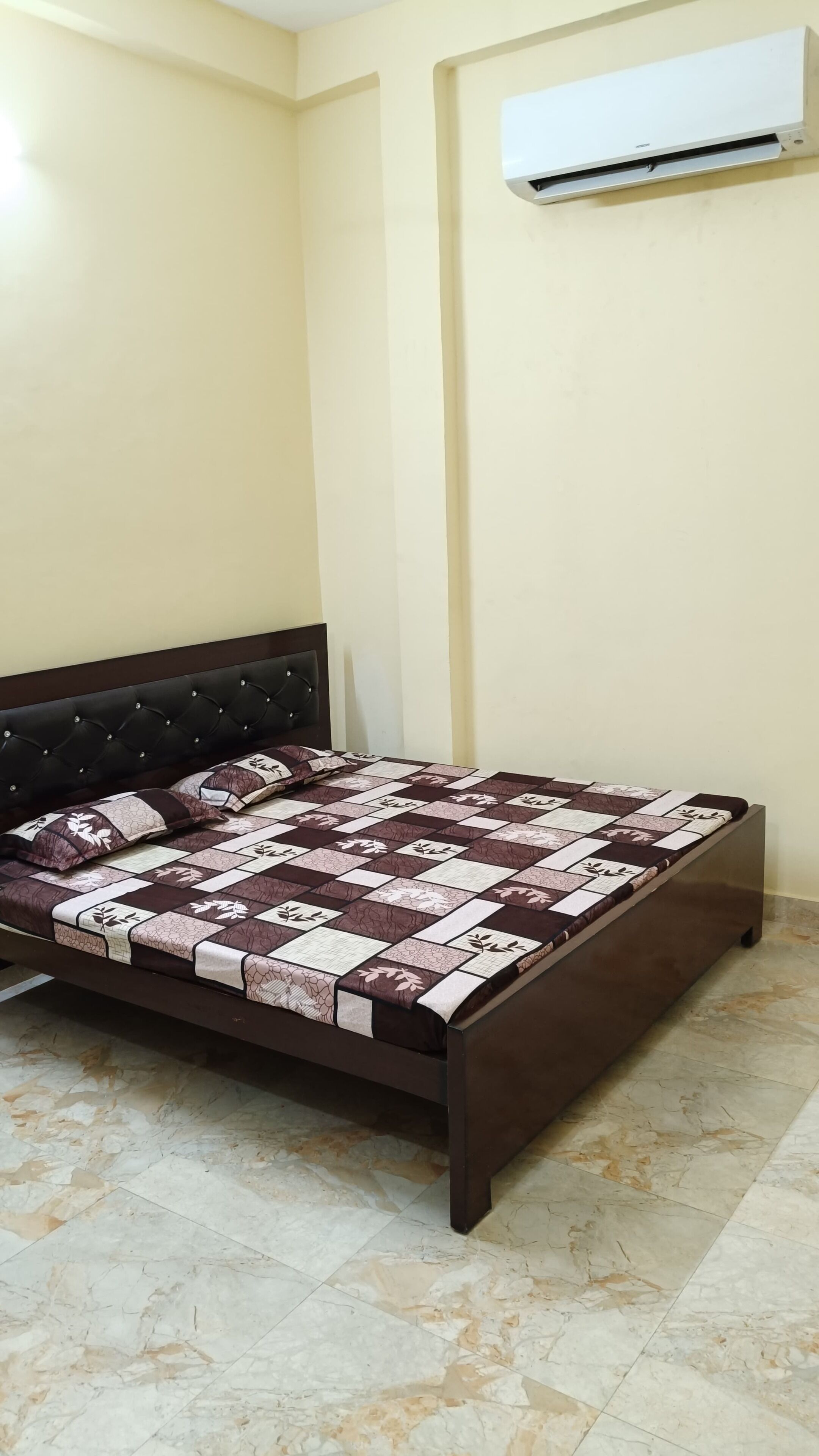 Un dormitorio con cama, cabecero oscuro, colcha estampada y aire acondicionado de pared.