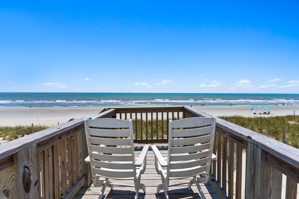 A wooden deck with two white chairs facing a beach.