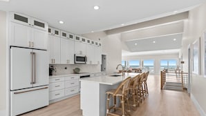 A spacious kitchen with white cabinetry, a large island, and a dining area with wicker chairs. - Chasing Sunrises | Newly Rebuilt Luxury Oceanfront Retreat (Ocean Isle Beach)