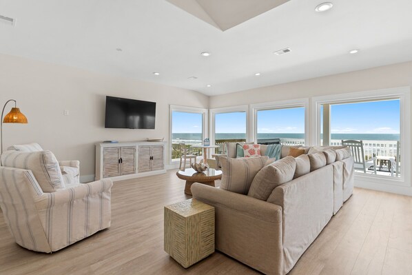 A spacious living room with a large sofa, a television, and a view of the sea.