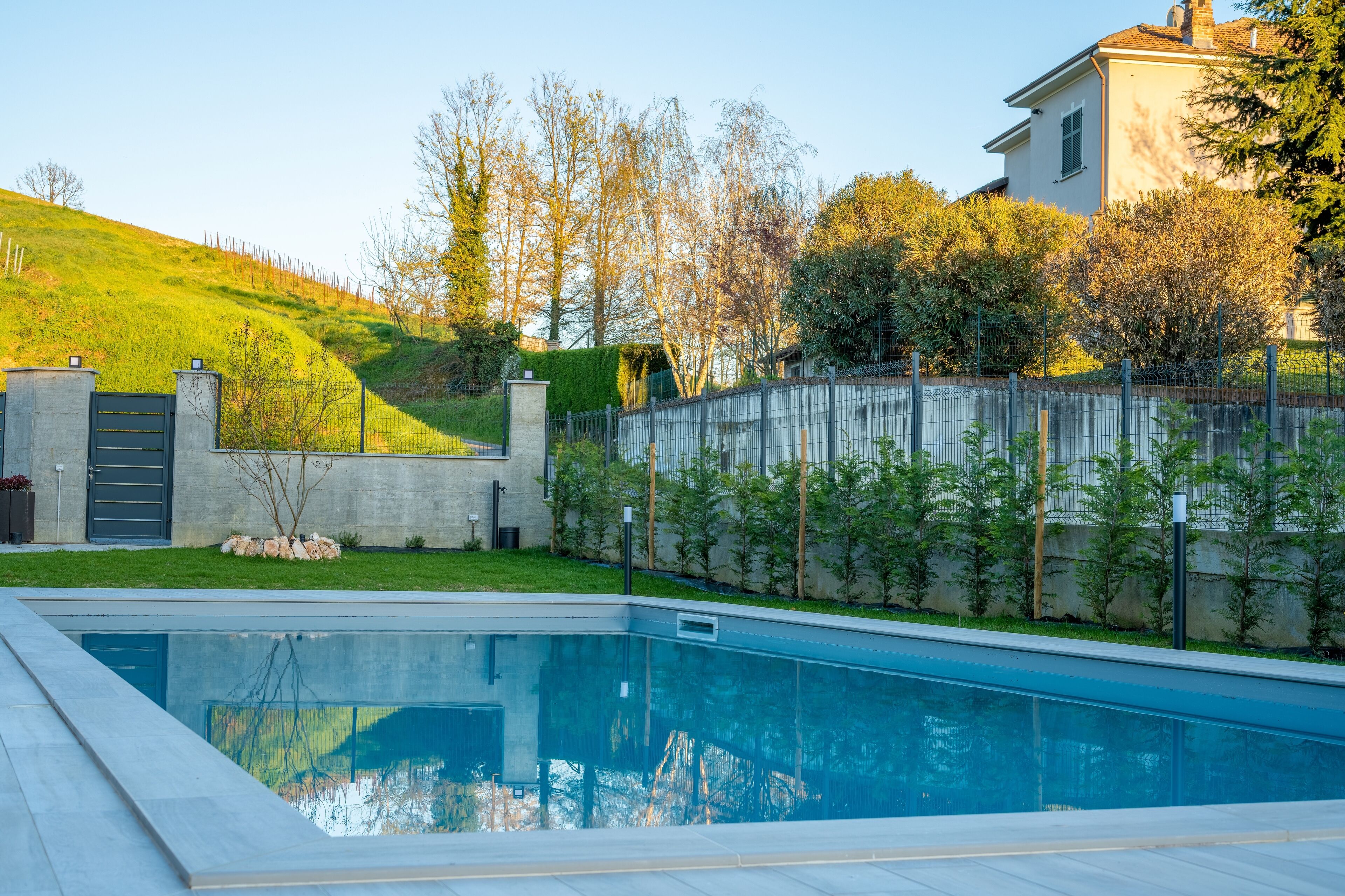 A swimming pool with a concrete deck, surrounded by a low stone wall and a metal fence.