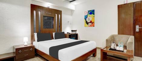 A hotel room with a wooden bed, a nightstand, a desk with a chair, a painting on the wall, and a wardrobe.
