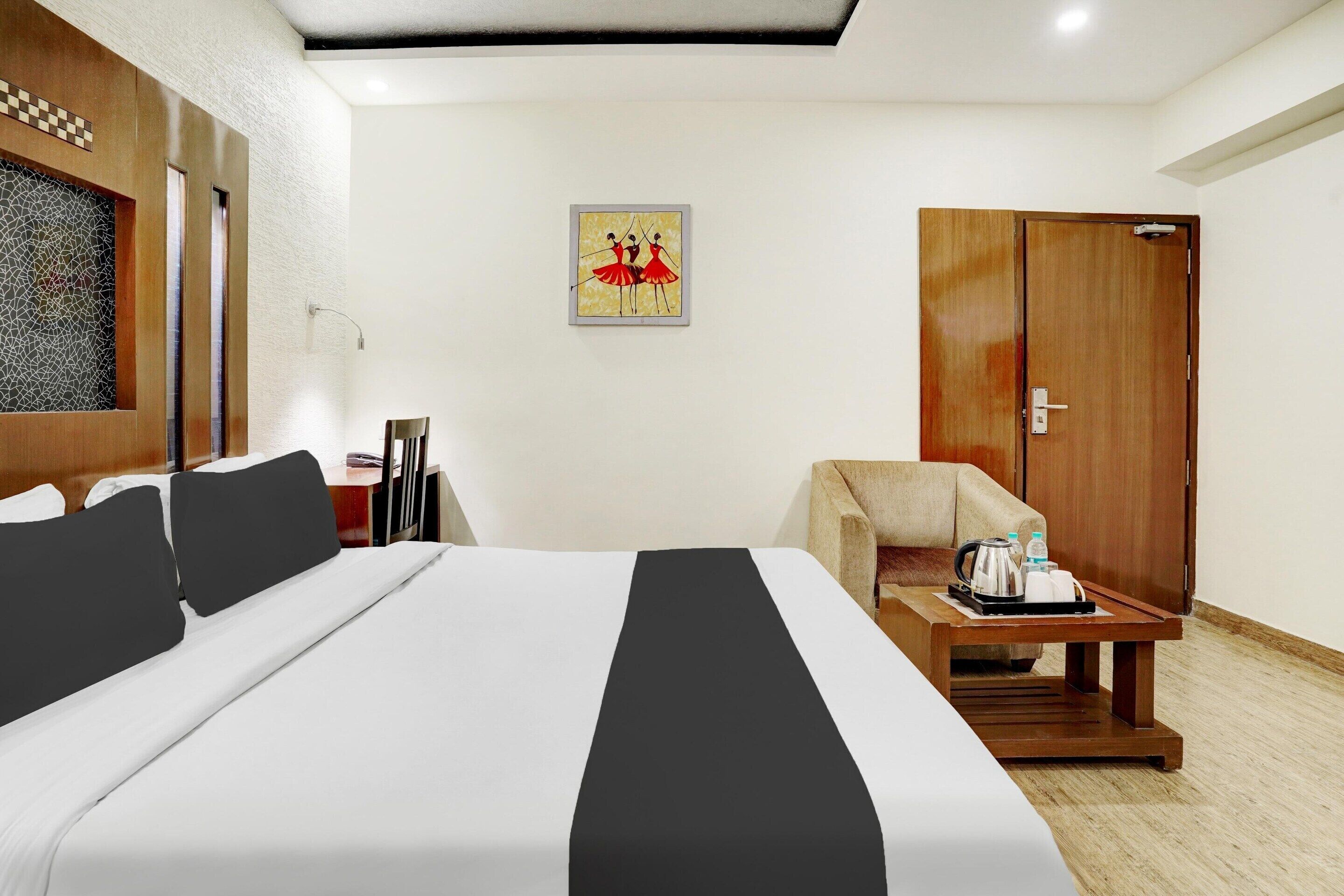 A hotel room with a bed, a chair, a small table with a tray, a wooden wardrobe, and a wall-mounted picture.