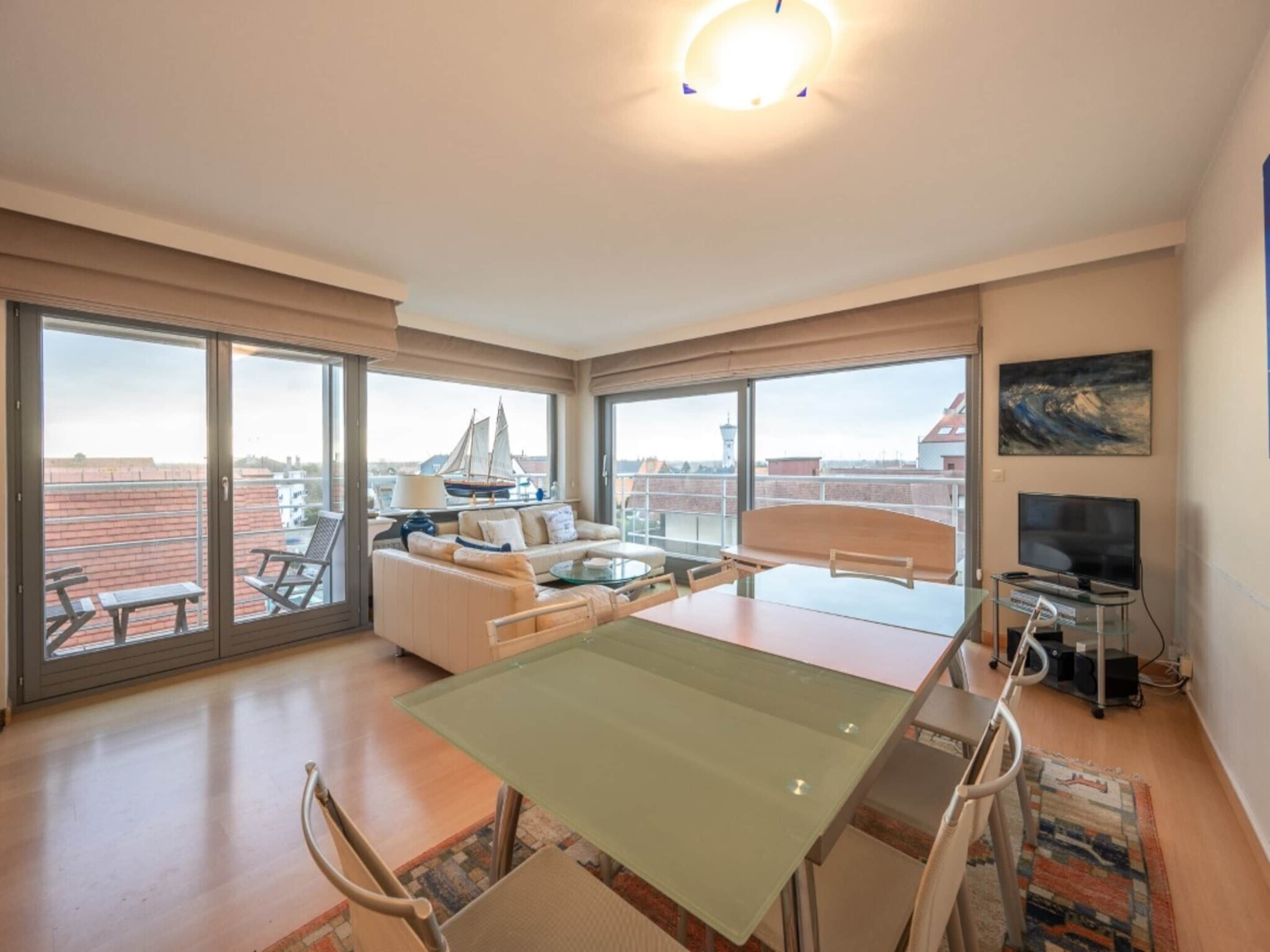 Un balcón amplio con vistas al horizonte de la ciudad, una zona de comedor con mesa de vidrio y un televisor a la derecha.