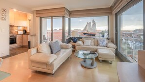A modern living room with a sofa, armchairs, a glass coffee table, and a view of a sailboat through large windows. - Apartment in Knokke-heist With Sea Views (Knokke-Heist)