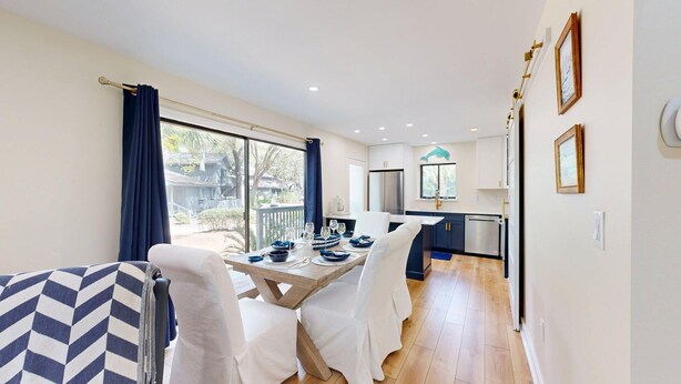Una cocina moderna con gabinetes blancos y electrodomésticos de acero inoxidable, un comedor con mesa de madera y sillas blancas, y vistas al patio trasero a través de ventanas amplias.