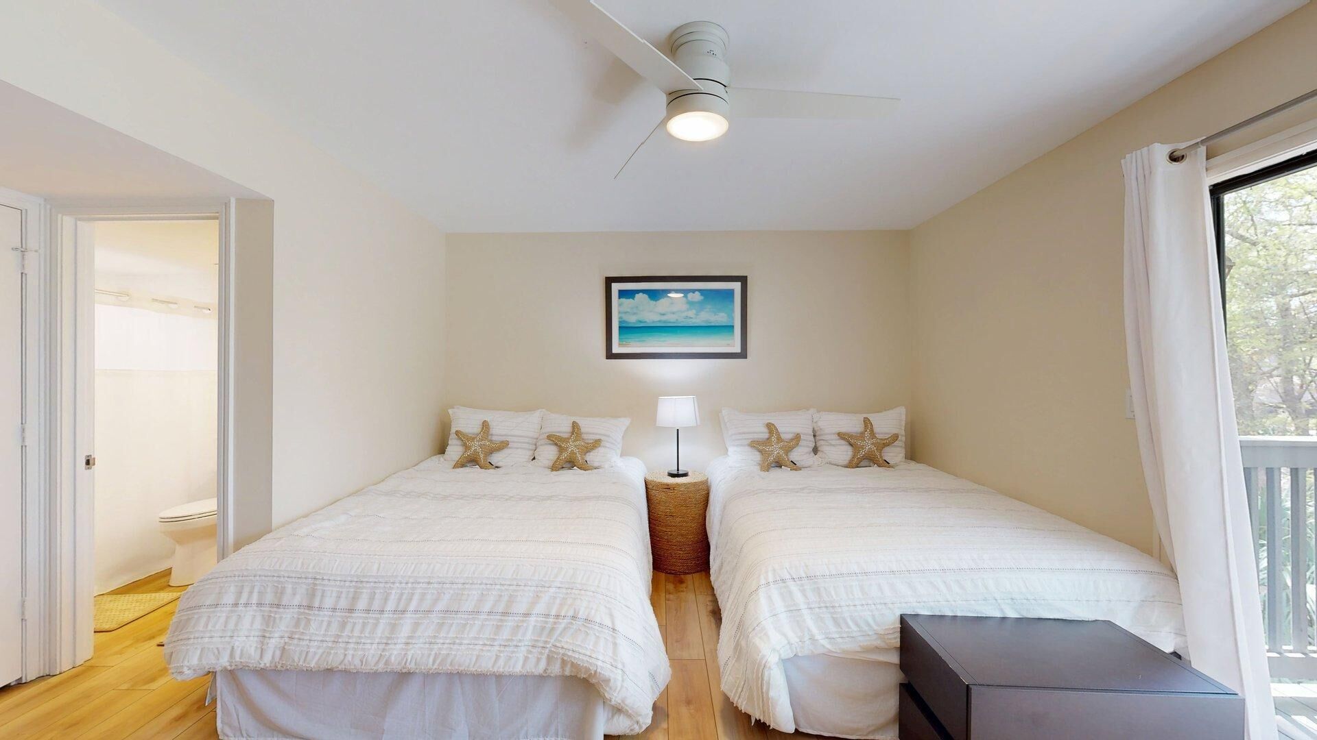 Une chambre avec deux lits, un ventilateur de plafond, un tableau encadré et une fenêtre avec des rideaux.