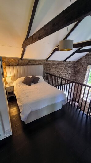 Een slaapkamer met een bed, een nachtkastje, een lamp, een stenen muur en een houten plafondbalk.