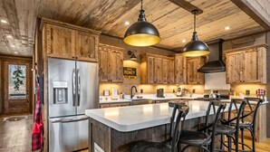 Eine rustikale Küche mit Holzschränken, einem Edelstahlkühlschrank, einer großen Kochinsel mit Barhocker und Pendelleuchten.