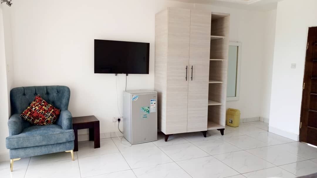 A living room with a blue armchair, a small table, a TV mounted on the wall, a wardrobe, and a trash bin.