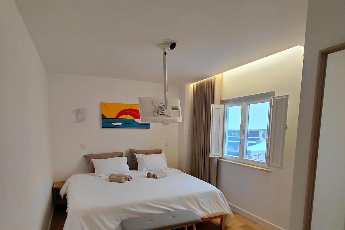 A bedroom with a bed, a bench, a ceiling-mounted projector, a window with curtains, and a wall-mounted artwork.