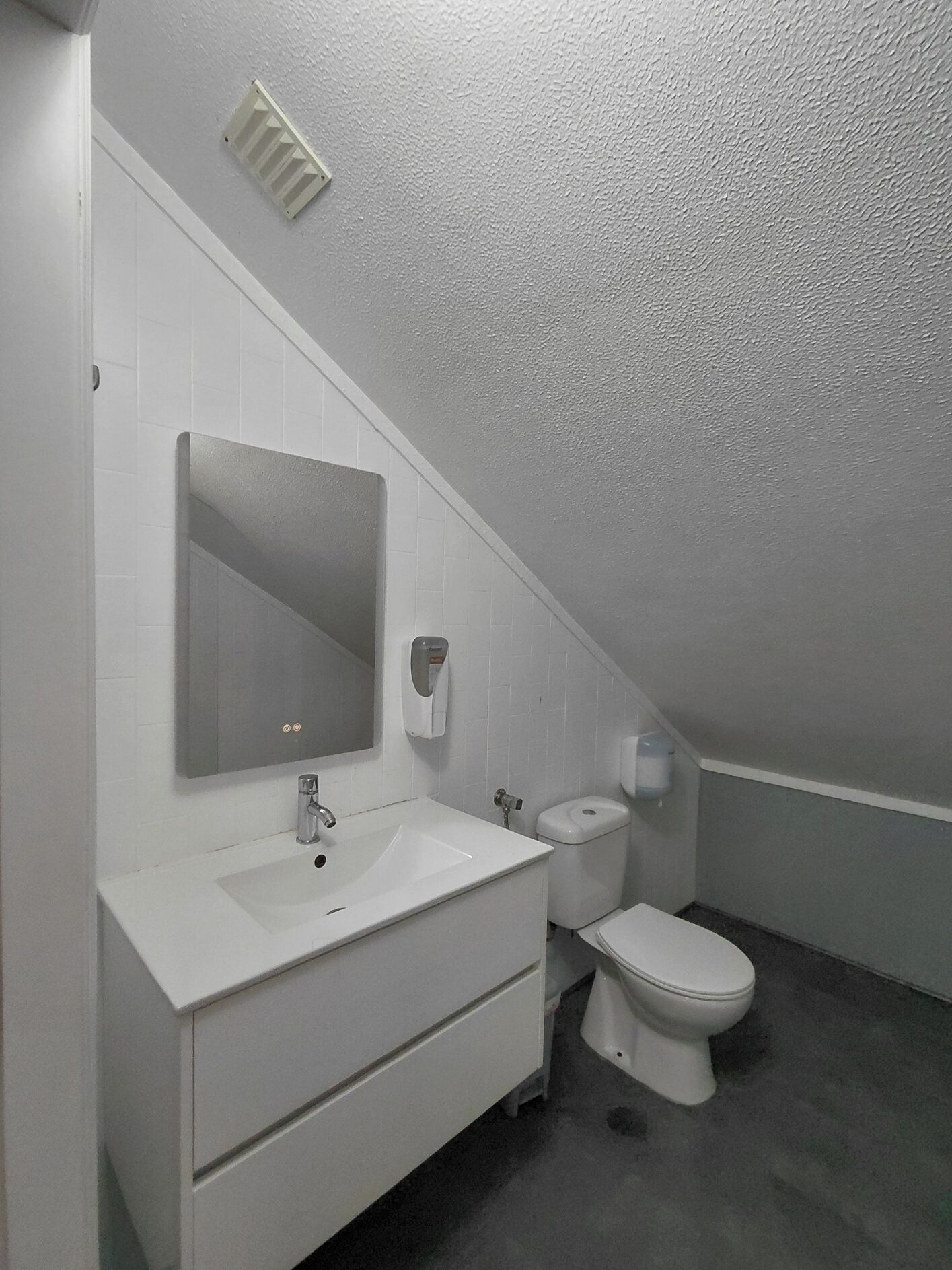 Un baño pequeño y moderno con un lavabo blanco, un espejo, un inodoro y una rejilla de ventilación en el techo.