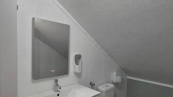 Un baño pequeño y moderno con un lavabo blanco, un espejo, un inodoro y una rejilla de ventilación en el techo.