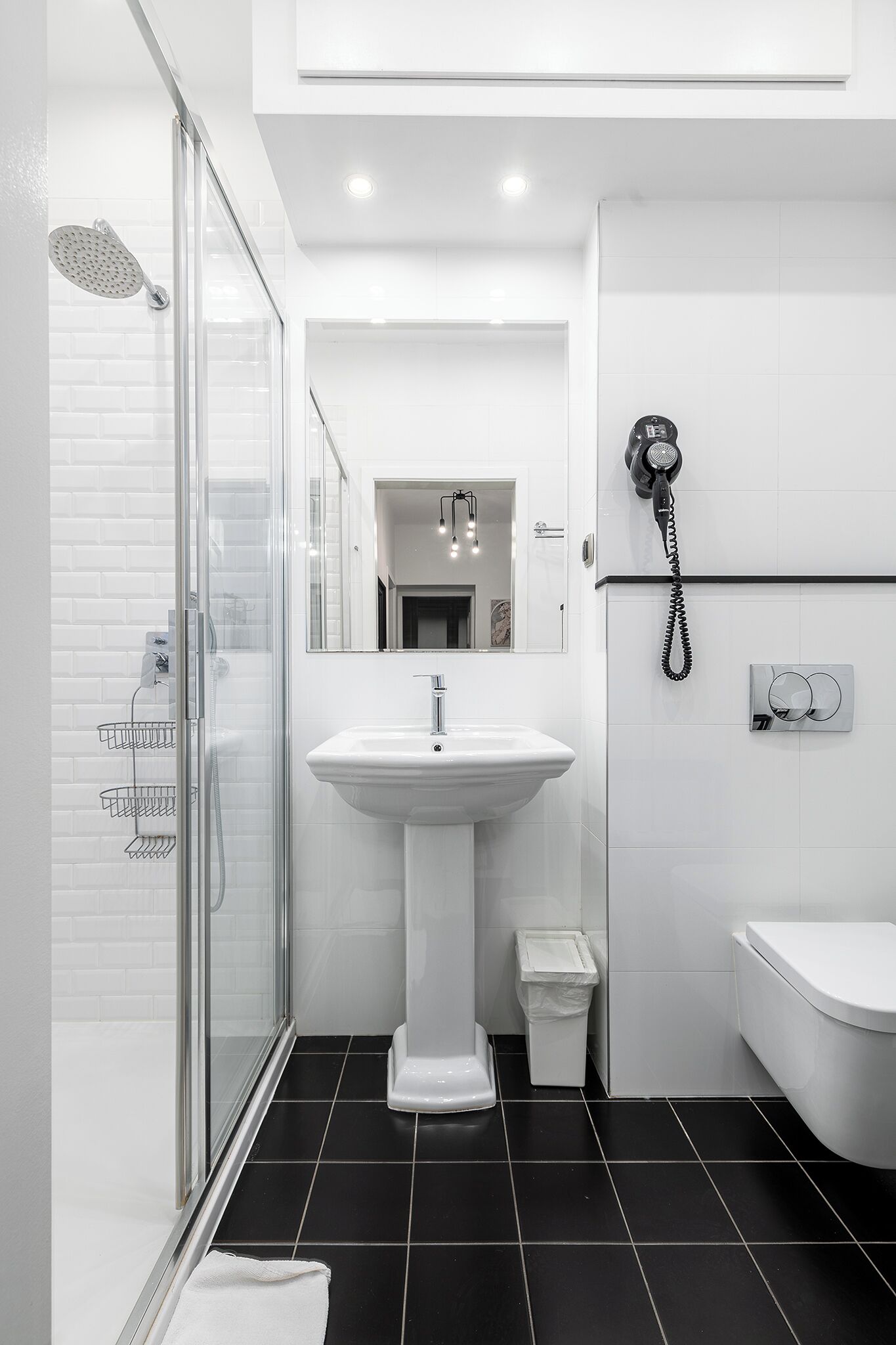 A modern bathroom with a glass shower enclosure, a pedestal sink, a toilet, and a hairdryer mounted on the wall.