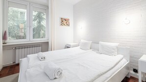 A modern hotel room with a large bed, white bedding, a radiator, a small bedside table, and a window with curtains. - RentPlanet - Apartament Chmielna (Warsaw)