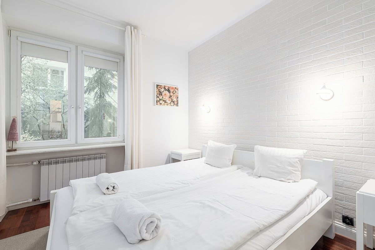 A modern hotel room with a large bed, white bedding, a radiator, a small bedside table, and a window with curtains.