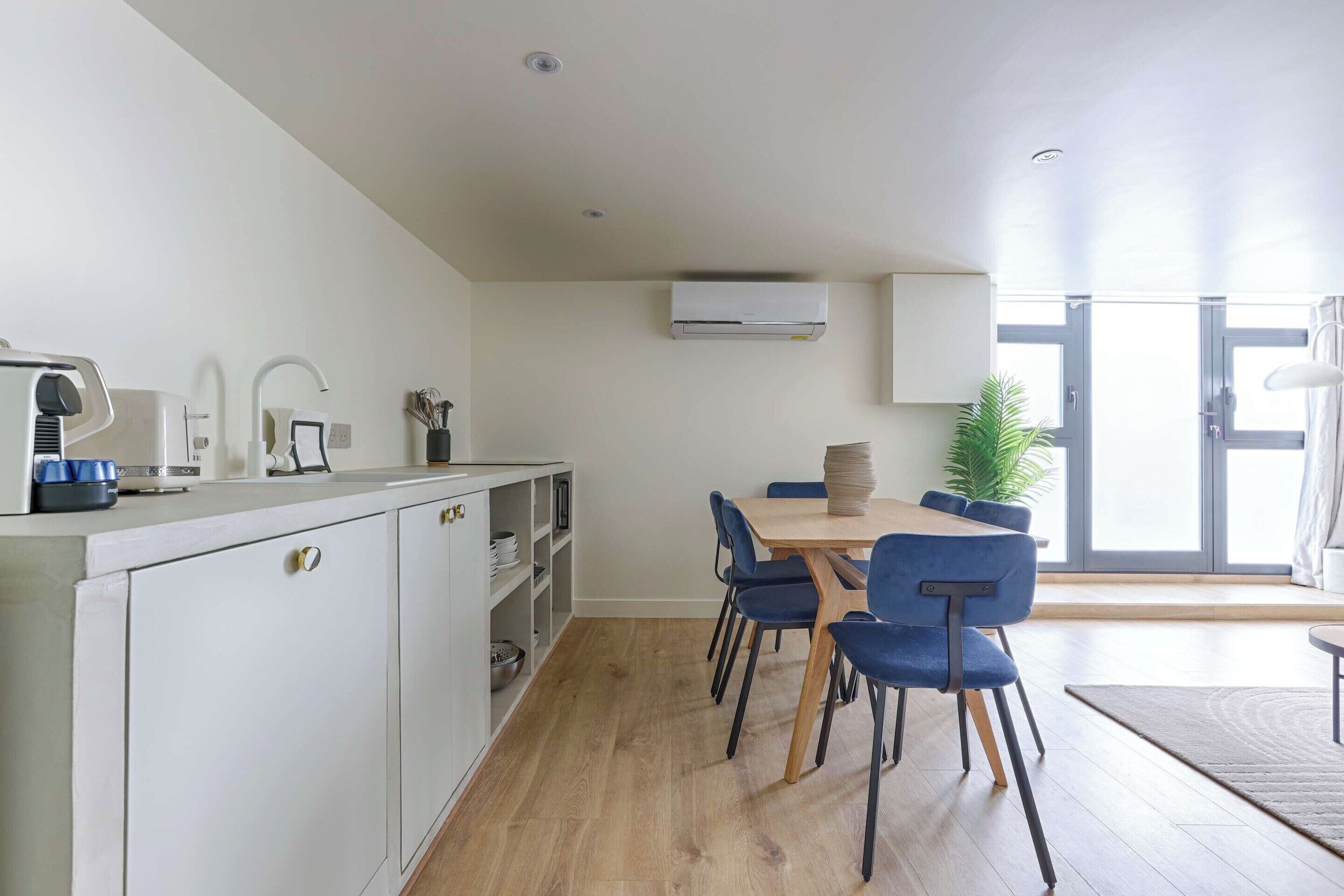 A modern kitchen with white cabinets, a wooden dining table with blue chairs, and a large window providing natural light.