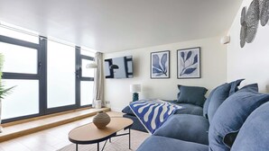 A modern living room with a sofa, a coffee table, and a TV. There are large windows and a plant. - Superb Air-conditioned Duplex - Bastille Le Marais (Paris)